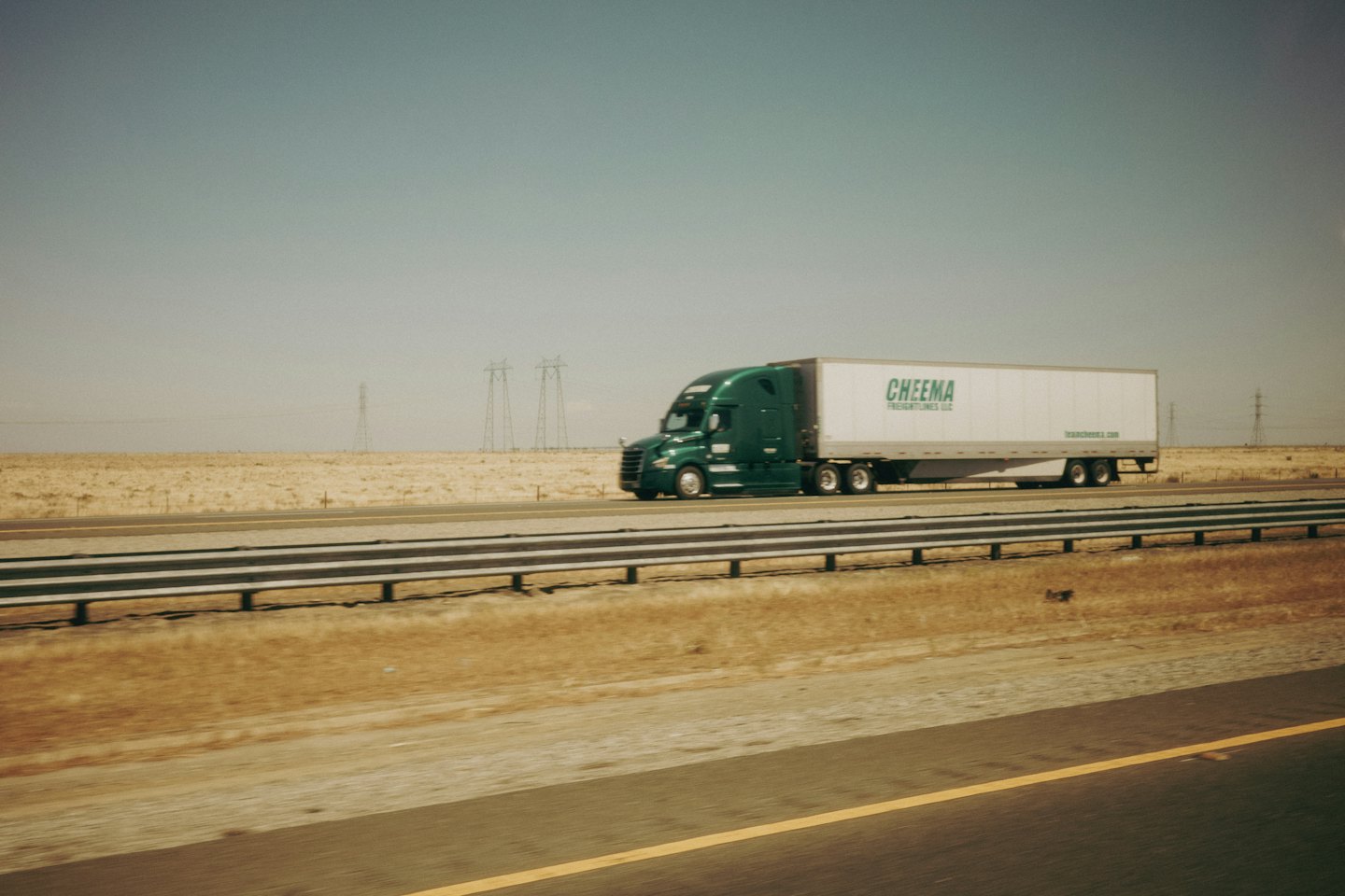 A green semi-truck drives down a highway next to a grassy field.